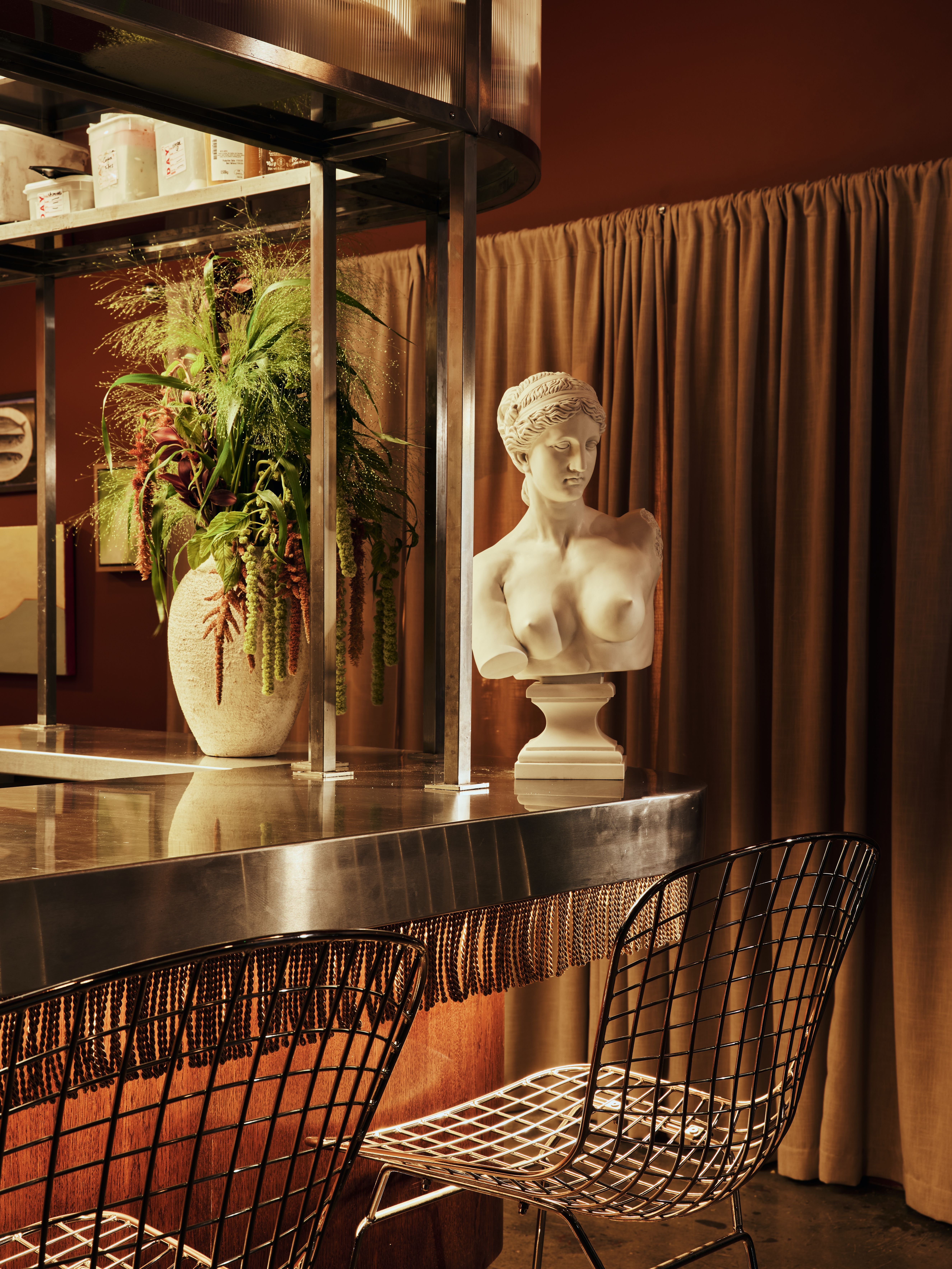 A swanky restaurant bar features brushed iron countertops with metallic fringes, a marble bust of a topless woman next to a wide ceramic vase filled with greenery, brown, suede curtains, and a cosmopolitan vibe.