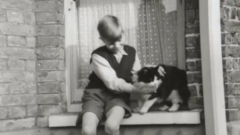 David Bowie Estate A black-and-white photograph of a young David Bowie sitting on a window sill, holding a black-and-white cat beside him, with brickwork and a door behind them.
