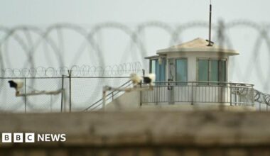 A view over a wall topped with a barbed wire, towards a guard tower, fences and CCTV security cameras