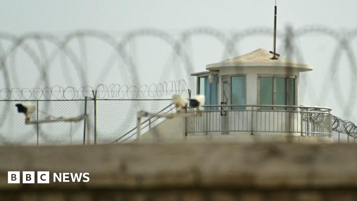 A view over a wall topped with a barbed wire, towards a guard tower, fences and CCTV security cameras