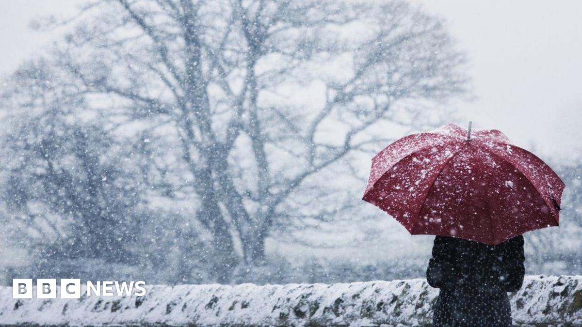 A woman wearing a long winter coat shelters under a red umbrella in a snow storm next to a snow-covered stone wall.