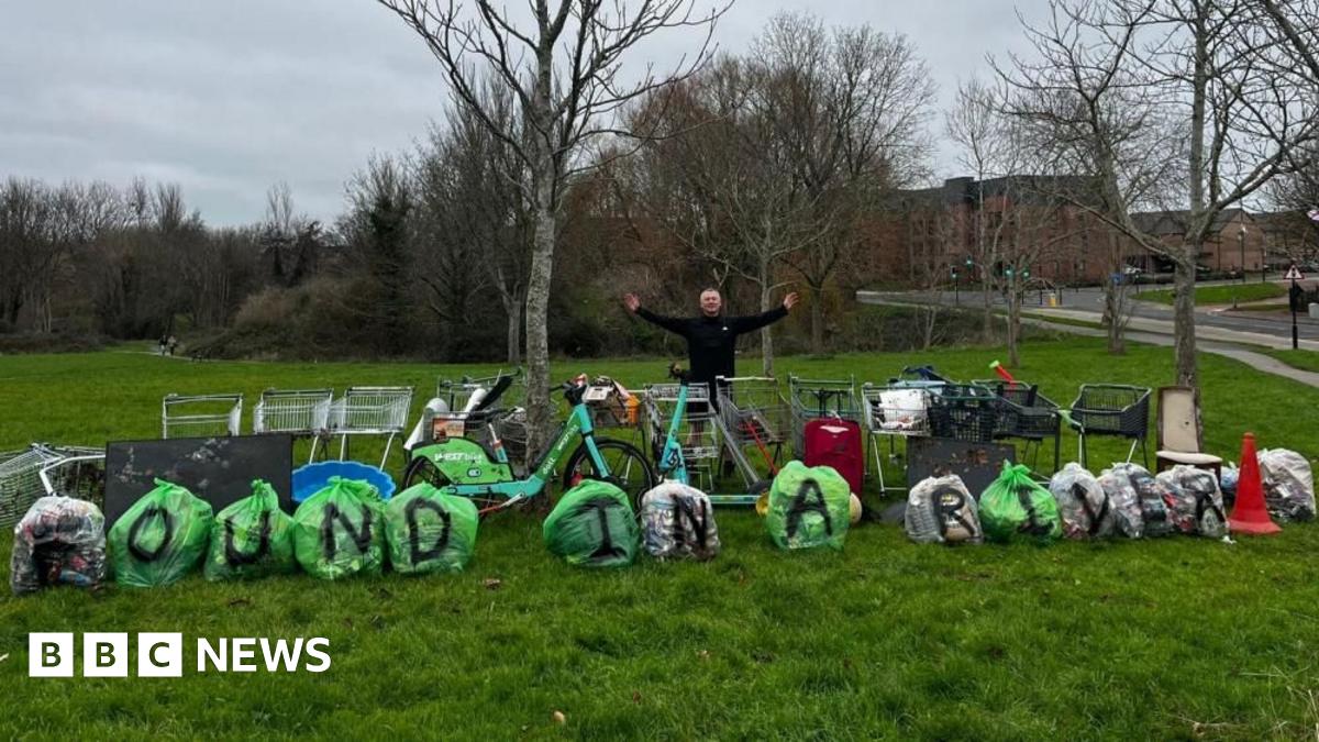 In a green park, Mark Rowlands poses with his arms out as he stands behind a row of rubbish backs with the letters 'FOUND IN A RIVER' written on them, as well as a row of shopping trolleys, e-bikes and e-scooters.