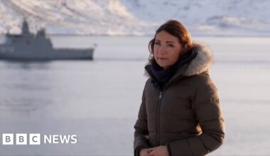 Katya Adler in a coat by the sea with a boat in the background.