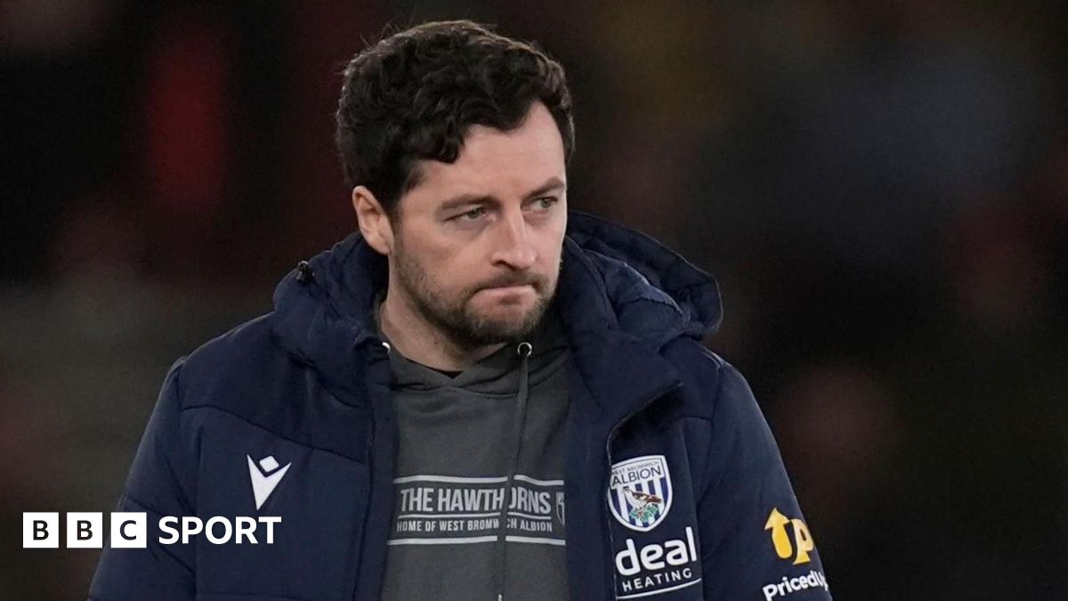 A man with dark hair and trimmed beard looks pensive at a football ground. He is wearing a dark tracksuit top