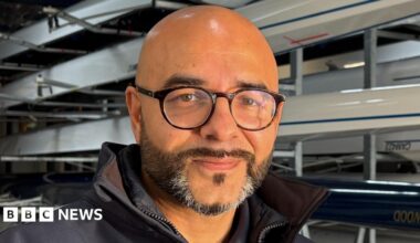 Omar Terywall, to the left of centre frame and looking directly out of the image. He is smiling slightly, has a dark, neatly trimmed beard, flecked with grey on his chin, and has on circular, black-framed glasses. He is bald. He is wearing a dark blue, collared top, with a white 'Cambridge Rowing' insignia on the right. Behind Omar are several sporting rowing boats mounted on racks inside a boathouse.