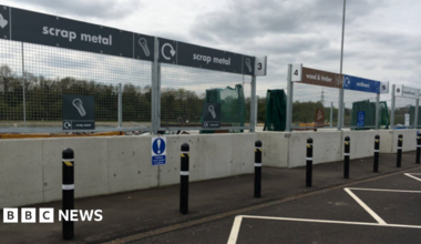 A household waste recycling centre with signs for scrap metal, wood and timber and cardboard. In front of the signs there are concrete barriers and a line of bollards.