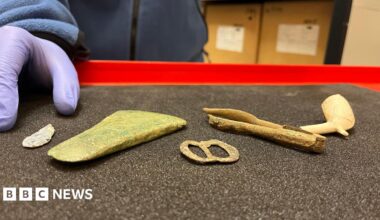 Two people wearing high-vis jackets and hard helmets crouched outside on an area of hard ground. One person is knelt with a hand inside a hole which has been dug into the ground.