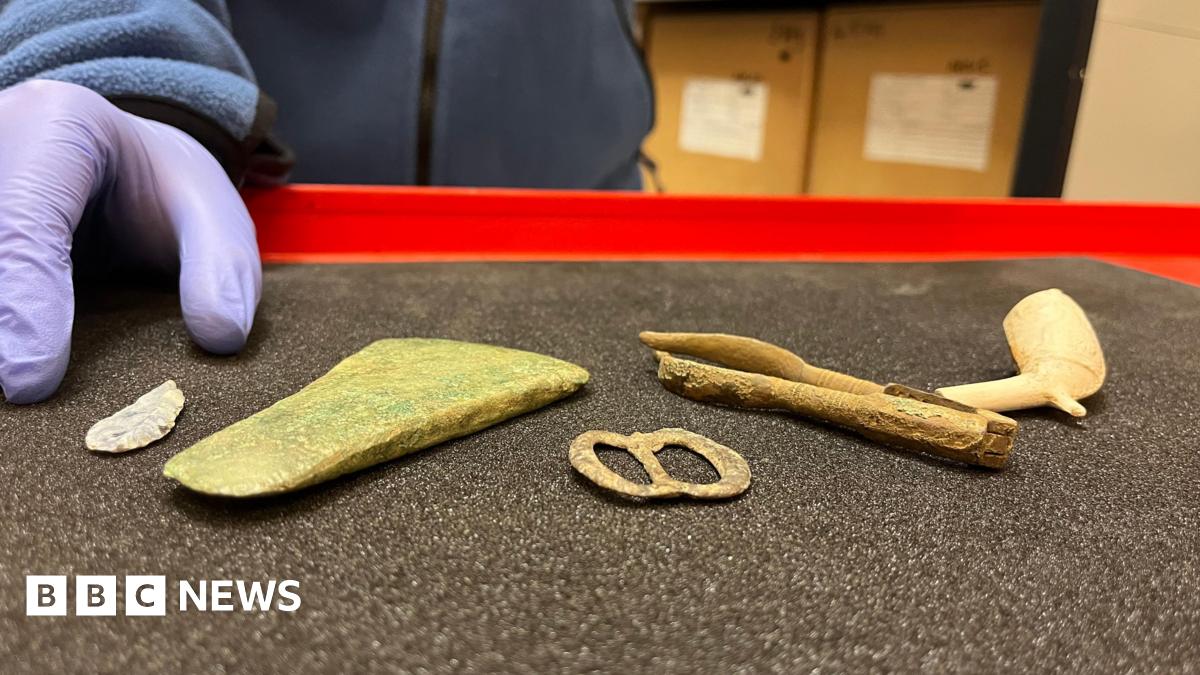 Two people wearing high-vis jackets and hard helmets crouched outside on an area of hard ground. One person is knelt with a hand inside a hole which has been dug into the ground.