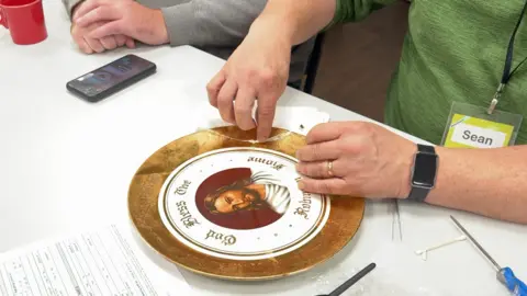 BBC news A family name plate is on the table. It has a wide gold rim. In the centre there is a depiction of Jesus with a red background. Golden writing surrounding Jesus says 'God Bless the Robinson Home'. A fixer wearing the name badge Sean has his hands on the plate, touching a large crack. He is wearing a green top.