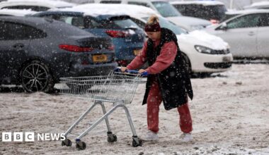 A woman pushes a trolley full of her supermarket shopping through far-stretching, thick brown slush in a car park. She wears a black sleeveless coat and red tracksuit. Her trolley is empty. In the background snow covered cars are visible