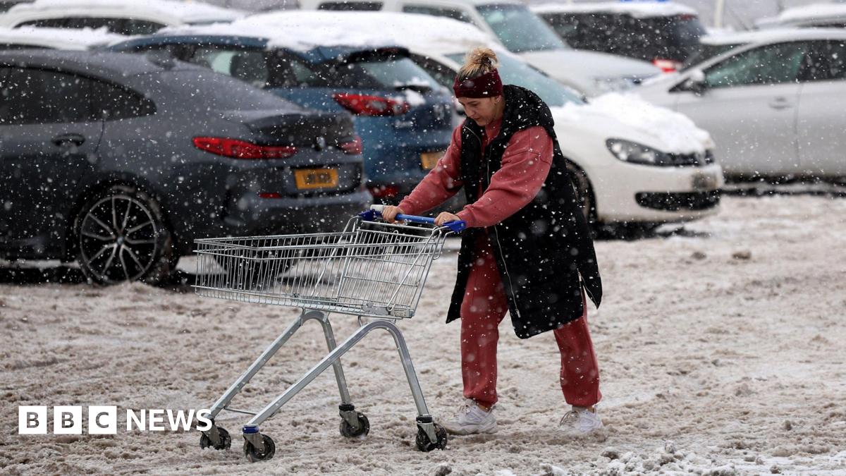 A woman pushes a trolley full of her supermarket shopping through far-stretching, thick brown slush in a car park. She wears a black sleeveless coat and red tracksuit. Her trolley is empty. In the background snow covered cars are visible