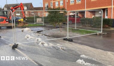 A street with houses down the far side and water running down the street with metal fencing closing off part of the street