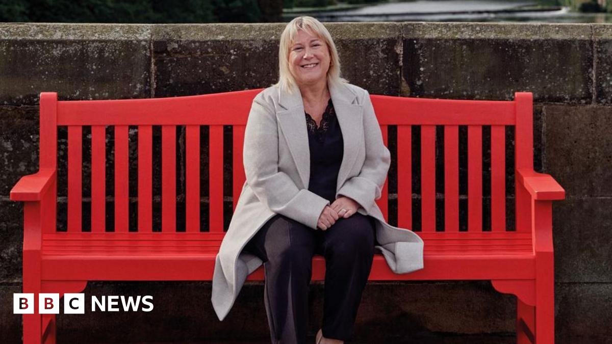Louise McGill sitting on a large red bench. She has shoulder-length blonde hair, is smiling and wearing a light grey long coat over a dark top and trousers. Behind her is a high stone wall.