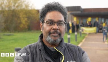 A man in a black jacket with grey sleeves with a yellow zip. He is stood outside, in the forefront of a protest, smiling at the camera