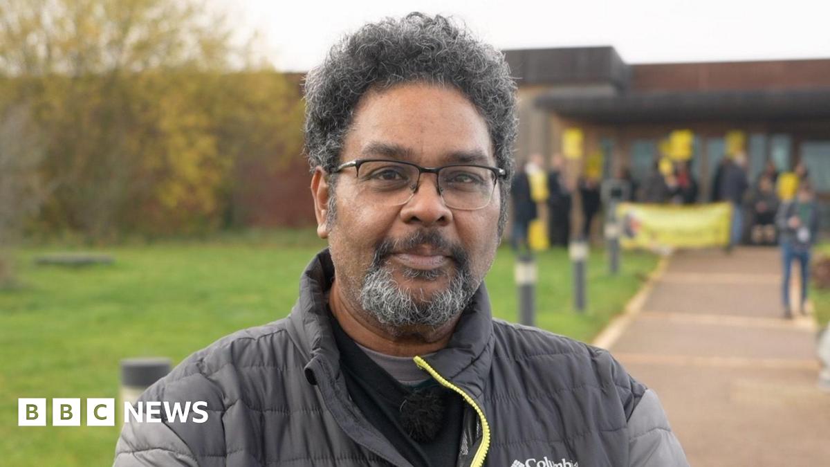 A man in a black jacket with grey sleeves with a yellow zip. He is stood outside, in the forefront of a protest, smiling at the camera