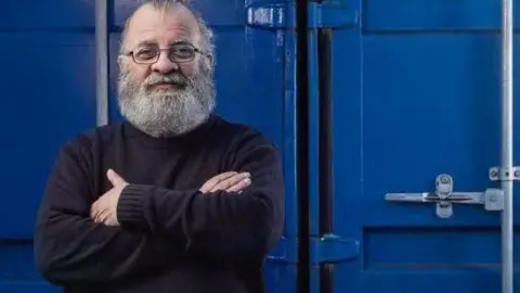 Ben Rafiqi Ben Rafiqi with his arms crossed and wearing glasses. He has a long grey beard and slick back balding grey hair. He is standing in front of a blue tank.