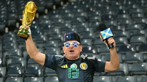Getty Images A man clad in Scotland gear holds a replica gold World Cup trophy above his head. He is wearing a tartan hat, Scotland strip and sunglasses with saltires on the lenses. He is standing in front of an empty blue stand. 