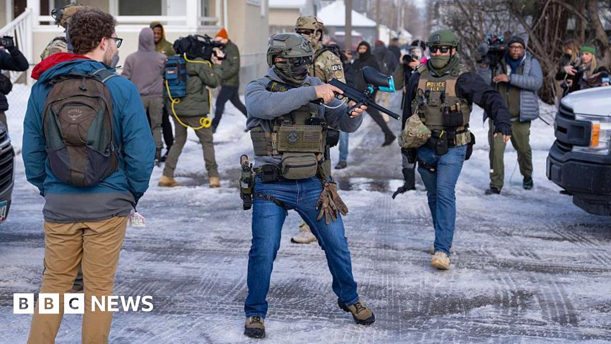 An officer with US Immigration and Customs Enforcement (right) and another federal officer holding a crowd control device (center) stand at a Minneapolis intersection where protesters had gathered after the death of Renee Good. Bystanders and journalists also stand in the intersection.