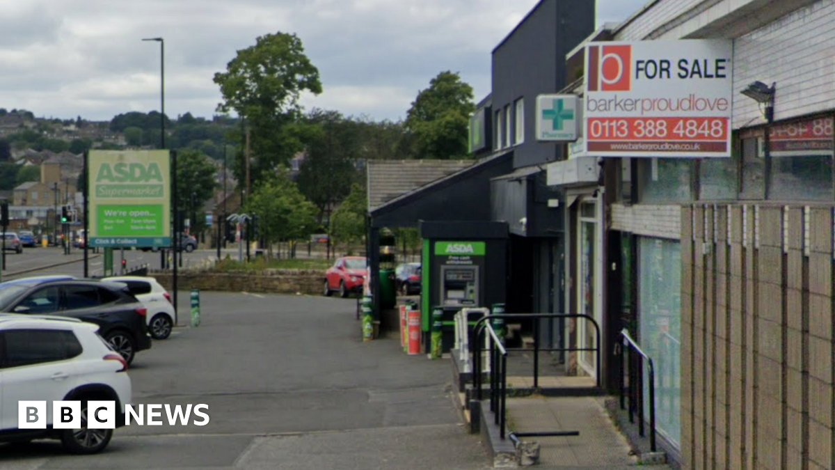 A strip of shops on a main road. In the foreground is a For Sale sign on an empty building. Behind is a pharmacy and an Asda.