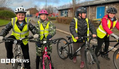 Four children are pictured on their bikes. They re all wearing high-vis jackets over their coats. A boy on the left is wearing a white helmet and smiling. A girl to his right is on a pink bike and wearing a pink and white spotted helmet. Two other boys are wearing black helmets.
