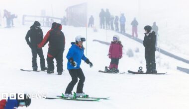 Skiers and snowboarders of various ages practise on a snowy slope in foggy conditions, with instructors and groups scattered across the background.
