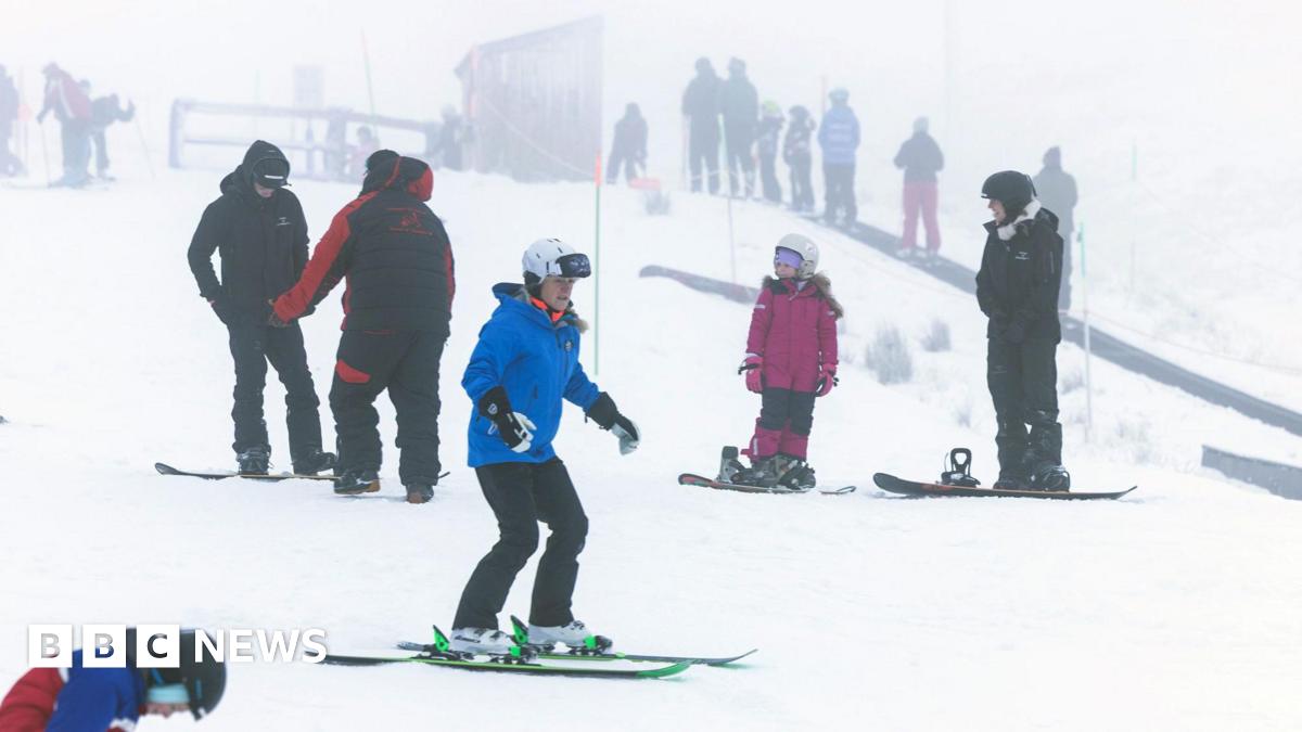 Skiers and snowboarders of various ages practise on a snowy slope in foggy conditions, with instructors and groups scattered across the background.