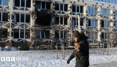 A man holding a dog walks near the site of a Russian drone strike on a private medical clinic. He has a hat and gloves and is walking on snow.