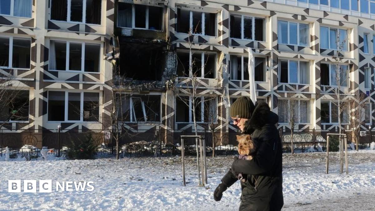 A man holding a dog walks near the site of a Russian drone strike on a private medical clinic. He has a hat and gloves and is walking on snow.