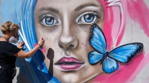 Upfest A woman paints a mural of a woman with pink and blue hair. The mural has a blue butterfly on it.