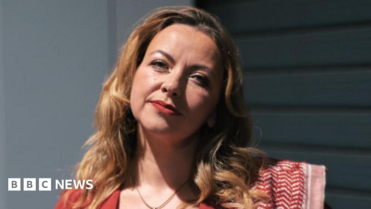 A close up head and shoulders shot of Charlotte Church. Her long brown wavy hair reaches her shoulder and she is wearing red lipstick. The top of her red dress, is visible, along with a necklace chain and a red scarf on one shoulder.