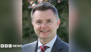 Graham Montgomery smiling at the camera. He is a man with short greying hair.  He is wearing a grey blazer, a pale shirt, and red tie with white and navy stripes.