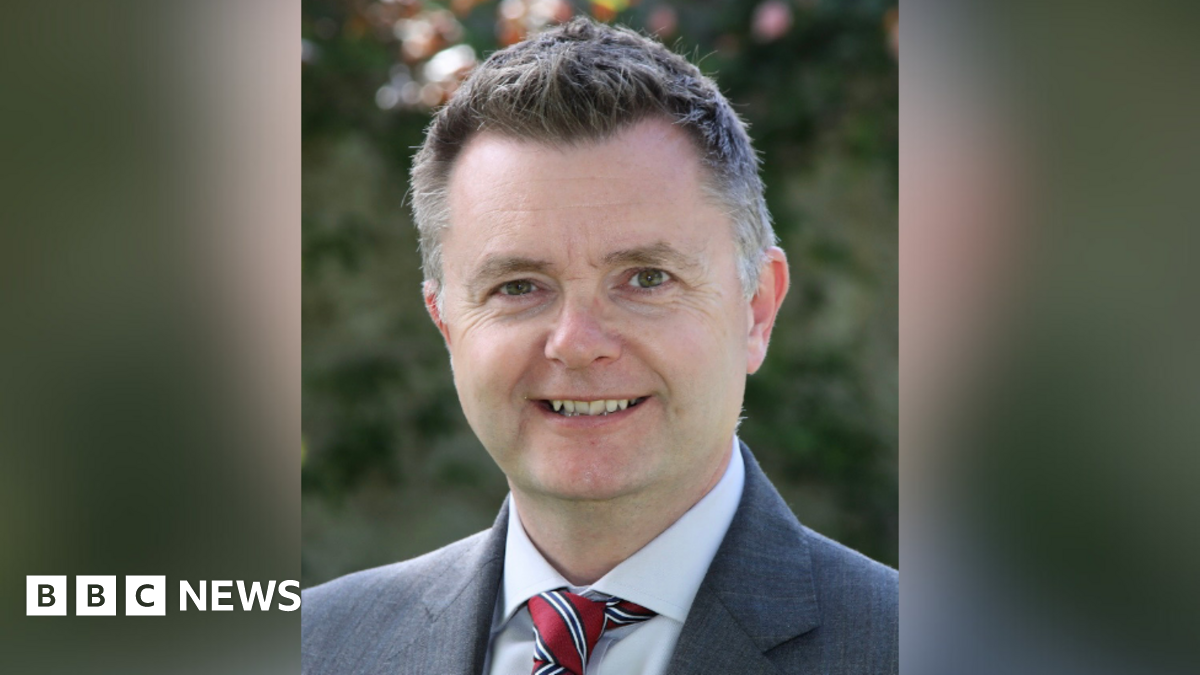 Graham Montgomery smiling at the camera. He is a man with short greying hair.  He is wearing a grey blazer, a pale shirt, and red tie with white and navy stripes.