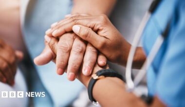 Millie Bobby Brown and David Harbour in character as Eleven and Jim Hopper. Eleven wears red shorts over grey leggings and a grey jumper. Jim Hopper wears a black leather jacket, a black beanie hat and dark trousers.They are speaking to director Frank Darabont on the set of Stranger Things. There is a vintage advertisement posted in the background with debrief on the ground.