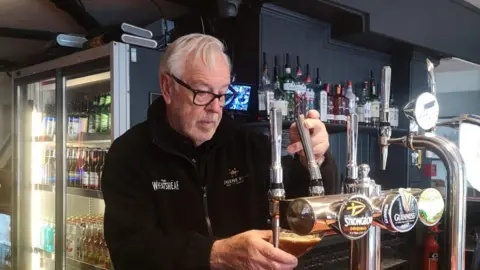 Geoff Robbins An older man in black fleece, with pub branding in white, is pulling a pint behind the bar in a pub. In the background there is a fridge with an internal light containing bottles of beer.