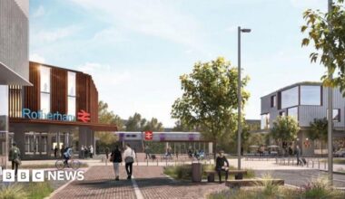 Miracle Moyo stands against a grey background, she is smiling and wears her long black hair in plaits held back with a black hair band.  A grey hoodie is visible under her cream coloured woolly fleece jacket.