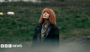 Kelly Reilly standing in a field, looking to the sky with her eyes closed. She has copper shoulder length hair with a fringe. She wears a green shirt and a green tartan coat.