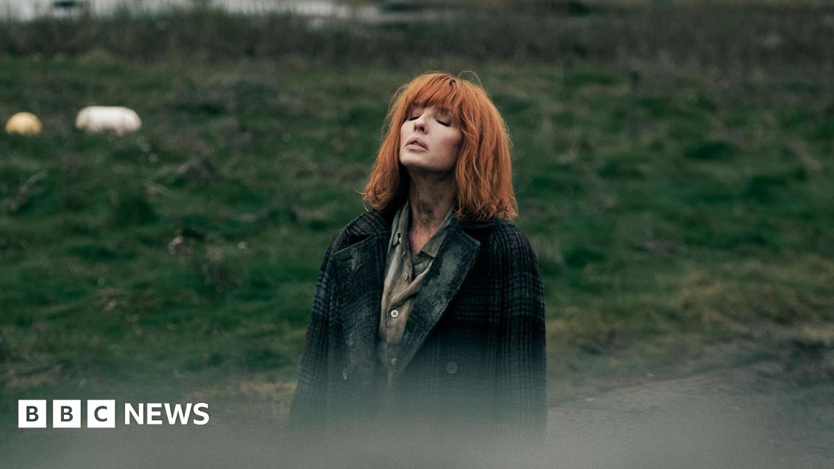 Kelly Reilly standing in a field, looking to the sky with her eyes closed. She has copper shoulder length hair with a fringe. She wears a green shirt and a green tartan coat.