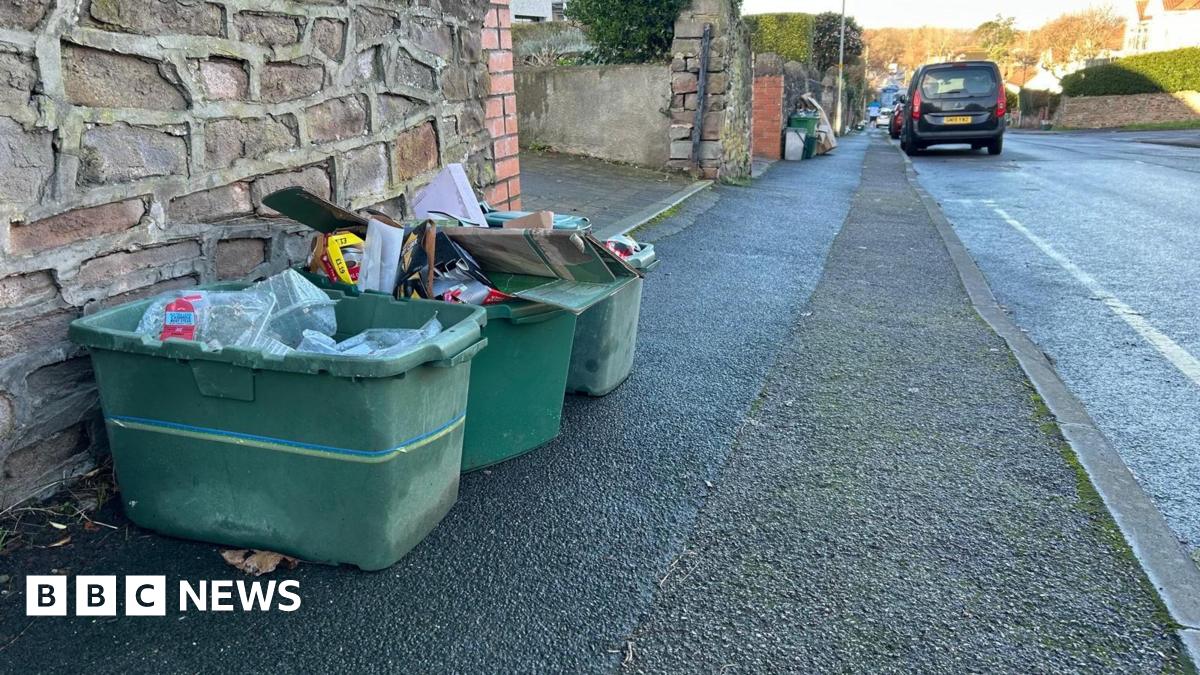 Three overflowing green recycling boxes sit on a pavement outside someone's front garden wall. They contain plastic packaging and cardboard.