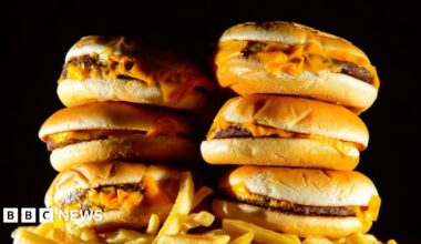 A stack of six cheeseburgers is piled on top of a portion of chips against a black backdrop.