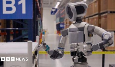 A silver and white Boston Dynamics humanoid robot lifting the lid of a container in a warehouse