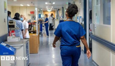 A woman wearing a blue NHS staff uniform walks through the corridor of a hospital away from us.