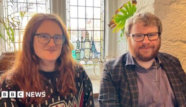 A woman with long ginger hair, black framed glasses and a blue patterned jumper next to a man with ginger hair, a ginger beard and black framed glasses in a blue shirt and blue chequed jacket in front of a stained glass window