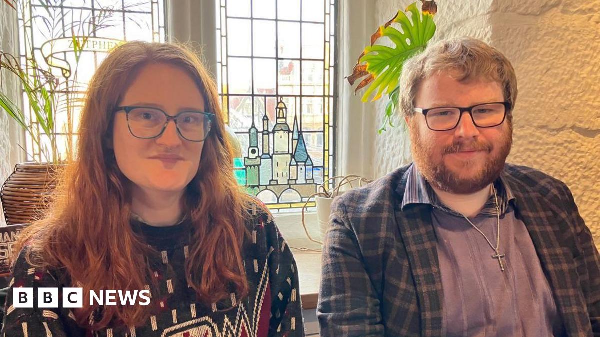A woman with long ginger hair, black framed glasses and a blue patterned jumper next to a man with ginger hair, a ginger beard and black framed glasses in a blue shirt and blue chequed jacket in front of a stained glass window