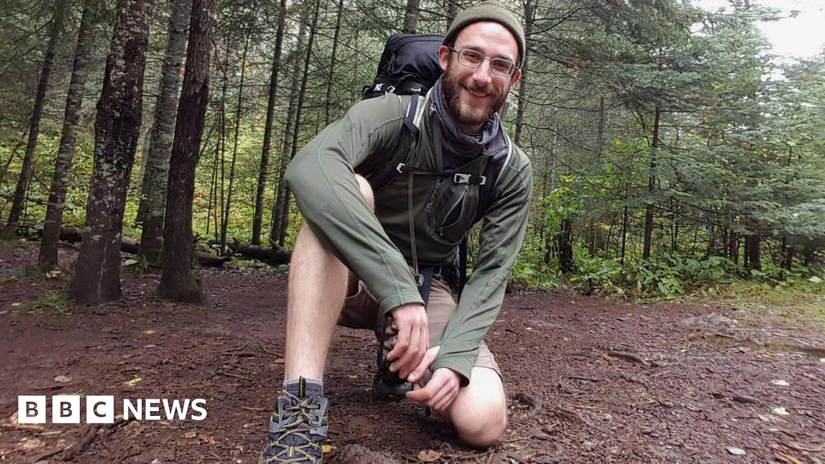 Alex is pictured in woodland wearing shorts and walking boots while kneeling on the ground