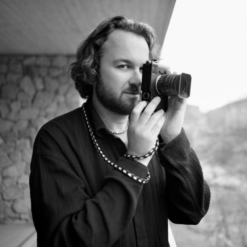 A bearded person with wavy hair holds a film camera up to their eye, ready to take a photo. They are standing on a balcony, with a stone wall and a blurred outdoor background behind them. The image is in black and white.