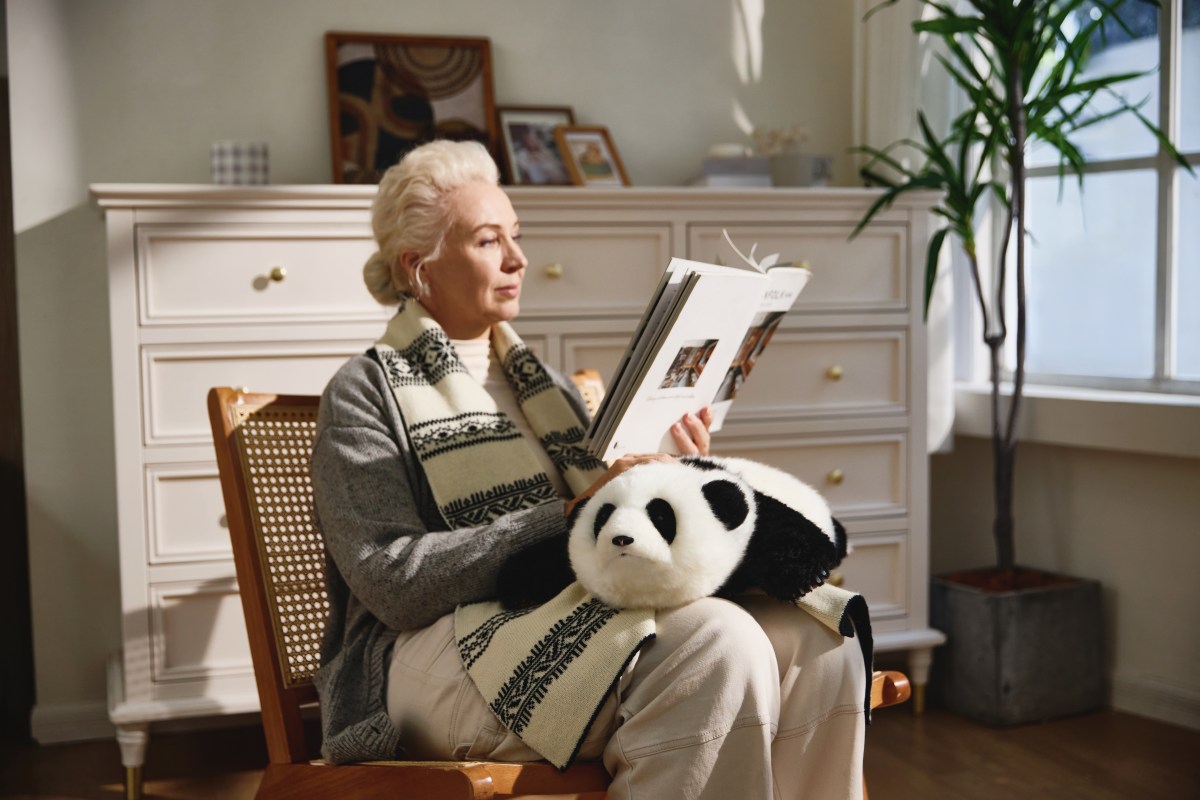 woman sits, reading, with a cuddly AI baby panda robot on her lap