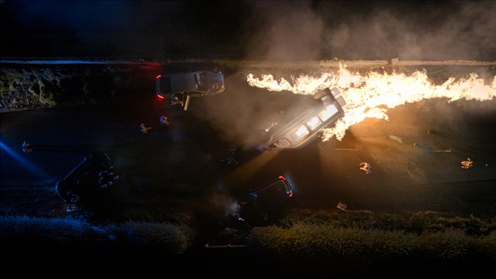 People stand amid several overturned cars and a trail of fire and smoke on a dark country lane in Corriedale