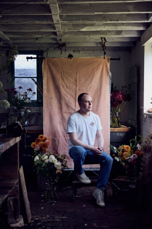 A man in a white t-shirt and jeans sits on a stool in a rustic room, surrounded by flower arrangements. A pink cloth hangs behind him, and natural light streams in from a window on the right.