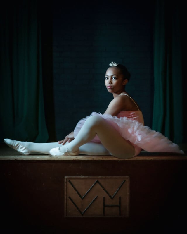 A ballerina in a pink tutu and tiara sits on a wooden stage with her legs extended and arms resting on her knees, softly lit against a dark background.