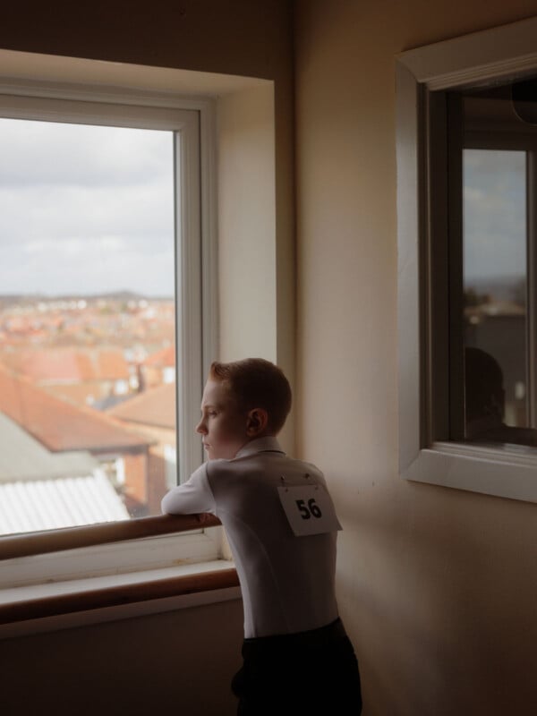 A boy wearing a numbered tag on his back stands by a window, looking outside with a thoughtful expression. Rooftops and a cloudy sky are visible through the window.